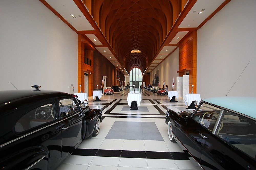 Classic black sedans parked on a polished tile floor in a long, top-lit car gallery hall